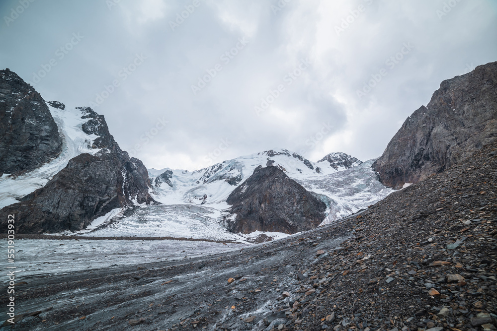 Fototapeta premium Dramatic landscape with two glacier icefalls on large snow mountain range with sharp rocks under gray cloudy sky. Long glacier with icefall in high altitude. Gloomy scenery in mountains in overcast.