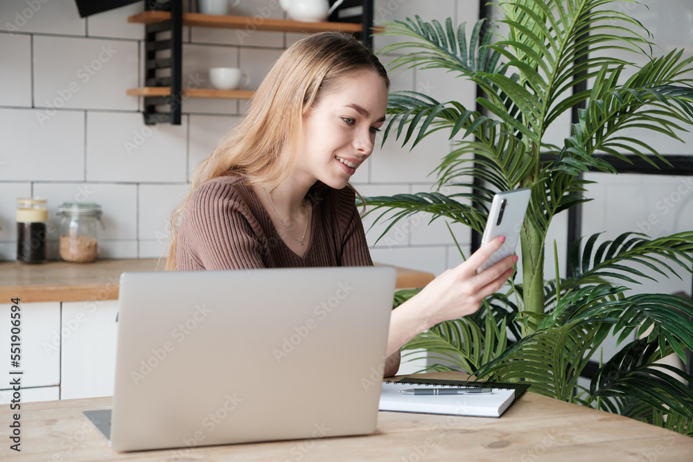 Beautiful smiling young female sitting at desk in kitchen using laptop computer and smartphone Beautiful smiling young female sitting at desk in kitchen using laptop computer and smartphone