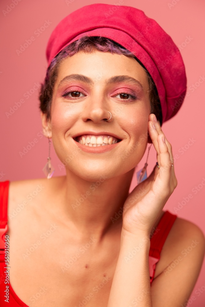 Young athletic woman with a short haircut and purple hair in a red top and a pink hat with an athletic figure smiles and grimaces looking at the camera on a pink background Young athletic woman with a short haircut and purple hair in a red top and a pink hat with an athletic figure smiles and grimaces looking at the camera on a pink background
