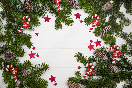 Green Christmas tree branches with pinecones decorated with red stars and Christmas candy canes on a white wooden background top view