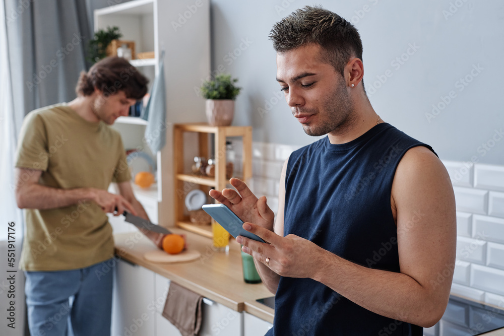 Same sex couple living together with focus on young man using smartphone in kitchen Same sex couple living together with focus on young man using smartphone in kitchen