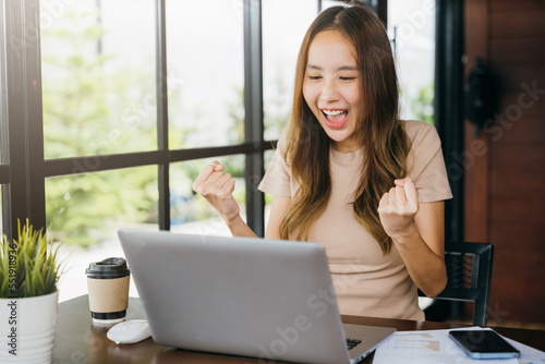 Excited happy Asian business young woman sitting with laptop computer at cafe desk raising hands up and celebrating success, female feeling winner rejoicing online win got new job at home