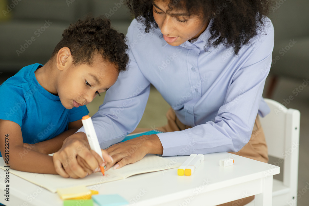 Female infant school teacher working one on one with a young schoolboy, sitting at a table writing in a classroom, front view Female infant school teacher working one on one with a young schoolboy, sitting at a table writing in a classroom, front view