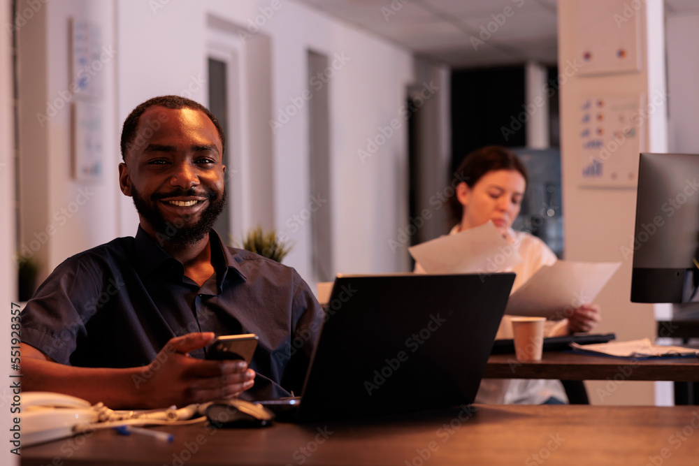 Smiling employee working on laptop, using smartphone app in coworking space portrait, project manager looking at camera at workplace desk. African american man writing report on computer