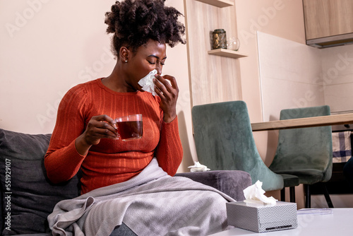 Woman sitting on a sofa, feeling sick. African American girl having flu, blowing her nose, and holding a cup of tea.