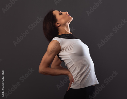 Strong sporty woman doing the arms and shoulders stretching arch exercise holding the hands in fist the low back to relaxing in sport white t-shirt wear on dark grey studio background. Closeup.