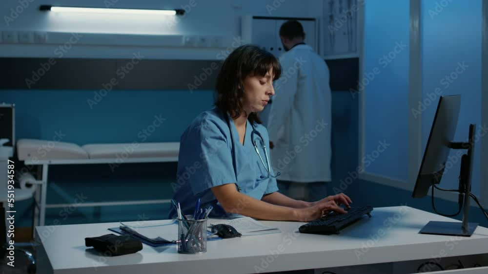 Physician nurse standing at desk table late at night in hospital office typing medical report during appointment. Assistant checking patient disease symptoms planning health care treatment Physician nurse standing at desk table late at night in hospital office typing medical report during appointment. Assistant checking patient disease symptoms planning health care treatment