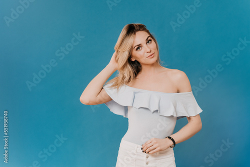 Gorgeous blonde wearing light blue blouse and white jeans, posing at studio, touching her hair, looking confident, being in good mood, over blue background with copy space. Happy people concept.