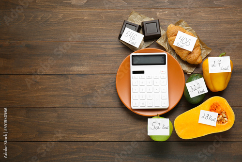 Flat lay composition of calculator and food products with calorific value tags on wooden table, space for text. Weight loss concept photo