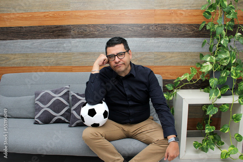 Latino adult man with glasses watches soccer game on TV on his sofa with his ball and gets angry and sad watching the football game