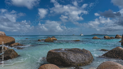 Picturesque granite boulders in the coastal waters of a tropical island. The waves of the turquoise ocean are beating against the rocks, foaming. The yacht is far away. Clouds in the blue sky. 