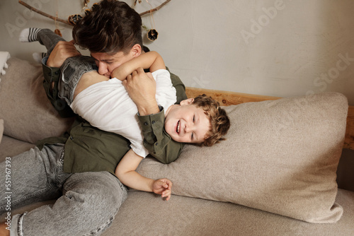 Young man sitting on sofa at home playing with his little son tickling him horizontal shot photo