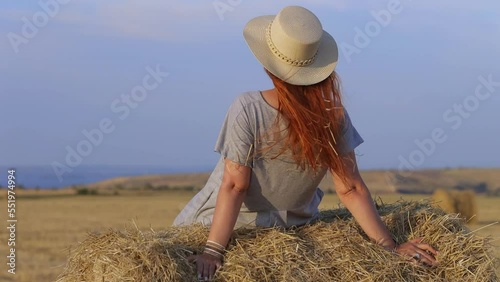 A beautiful young woman in a hat and a summer dress is sitting on a sheaf of hay in a field. Rural nature, wheat field, rest in the country, unity with nature