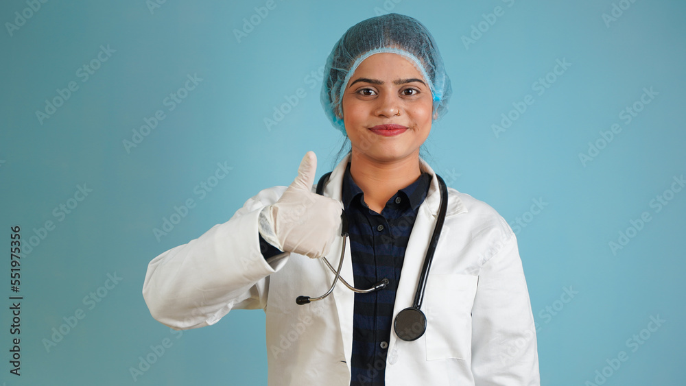Portrait of a young female doctor gesturing thumbs up, Cheerful Asian Indian woman doctor in apron and stethoscope isolated over blue studio background Portrait of a young female doctor gesturing thumbs up, Cheerful Asian Indian woman doctor in apron and stethoscope isolated over blue studio background