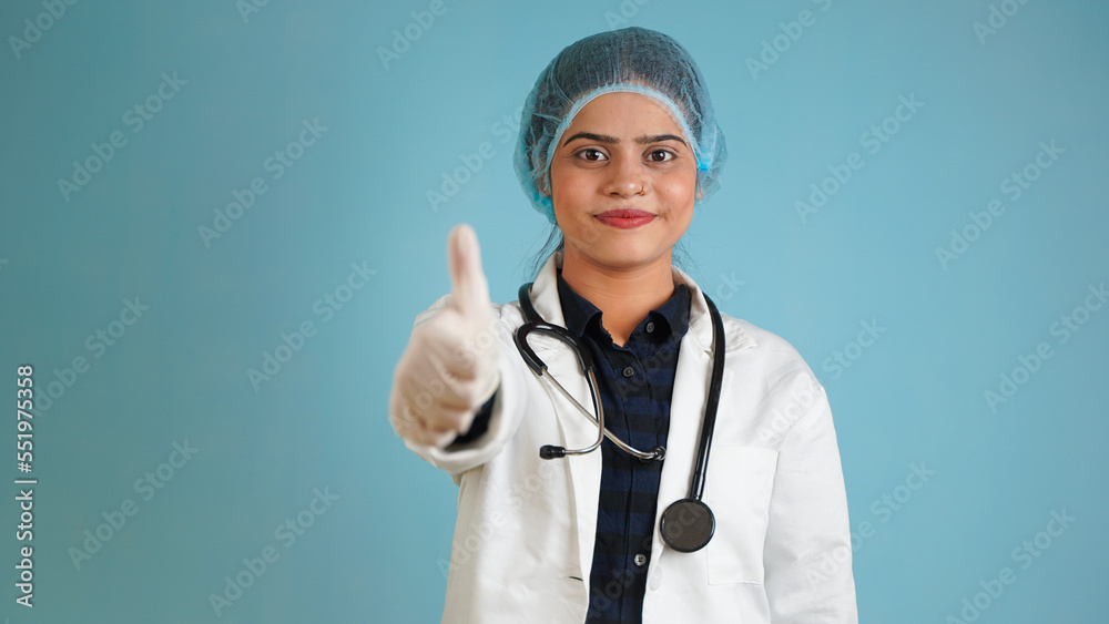 Portrait of a young female doctor gesturing thumbs up, Cheerful Asian Indian woman doctor in apron and stethoscope isolated over blue studio background Portrait of a young female doctor gesturing thumbs up, Cheerful Asian Indian woman doctor in apron and stethoscope isolated over blue studio background