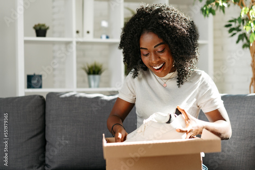 Young african woman sit on couch at home unpacking parcel cardboard box with online purchase photo