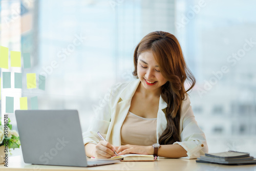 Young Asian businesswoman sitting happily working on her laptop and taking notes fluently and smiling happily at her assignment.