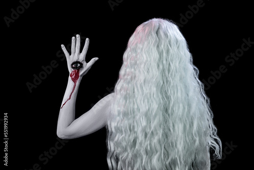 A woman in a white wig and an eye on her hand stands with her back on a black background