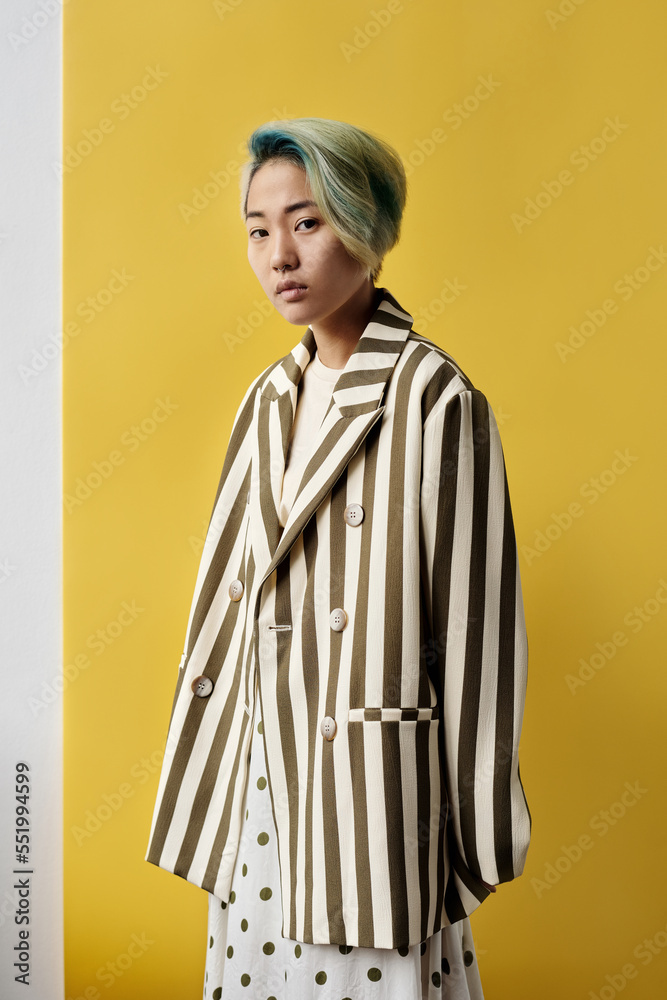 Medium long fashion shot of young Asian woman wearing stylish outfit standing against yellow wall background looking at camera
