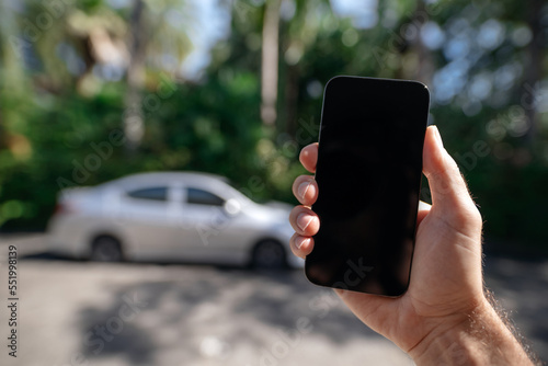 Smartphone mockup in hand against the background of a street and a car in summer. Car rental or car sharing.