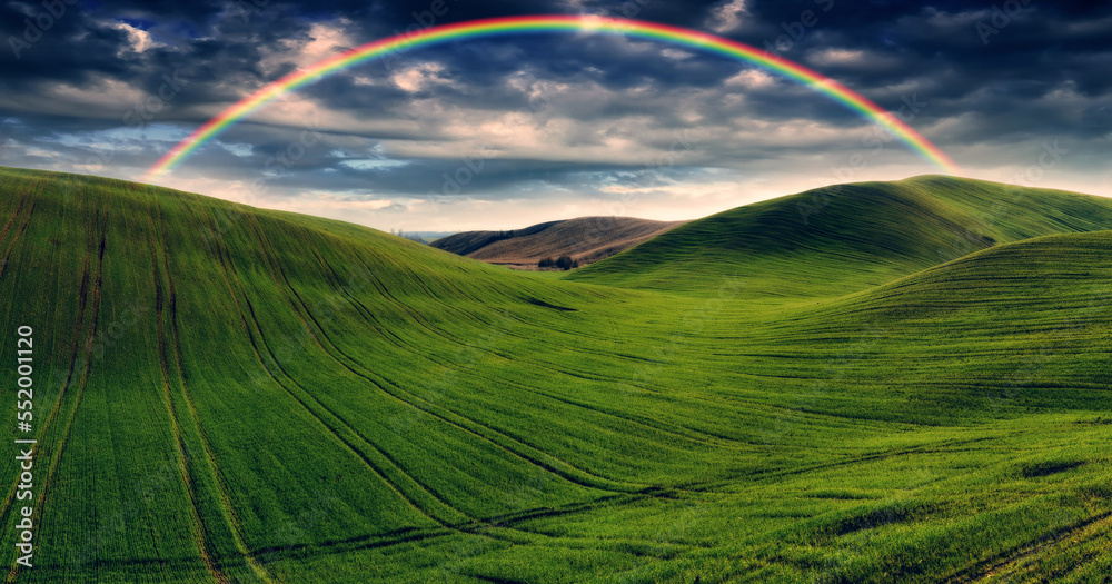 Obraz premium Scenic view of rainbow over green field. dramatic gray sky over a picturesque hilly field