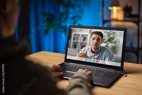View from shoulder of female sitting at desk during evening time and having video call on modern laptop. Concept of connection people.