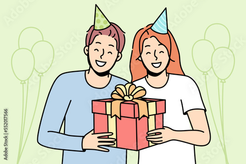 Guy, girl in party hats are smiling, holding gift box tied with ribbon. Meeting guests at holiday. Celebrating birthday, anniversary, significant event. Vector line art multicolored illustration.