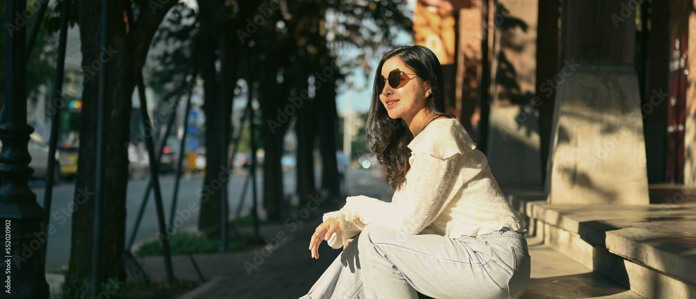 Fototapeta premium Modern millennial women in sunglasses sitting on stairs at the shopping district of a city. Banner background, panoramic view
