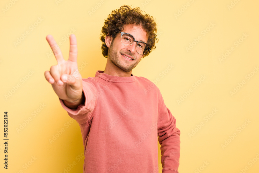 Young smart caucasian man on yellow background joyful and carefree showing a peace symbol with fingers. Young smart caucasian man on yellow background joyful and carefree showing a peace symbol with fingers.