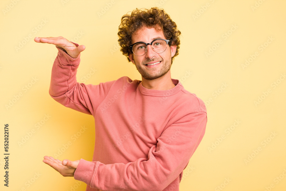 Young smart caucasian man on yellow background holding something little with forefingers, smiling and confident.