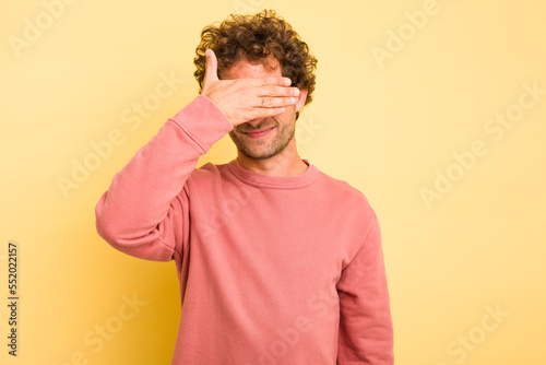 Young smart caucasian man on yellow background covers eyes with hands, smiles broadly waiting for a surprise. © Asier