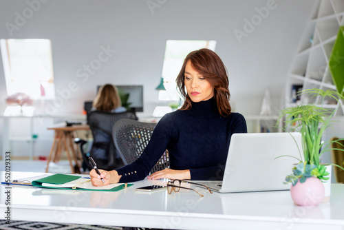 Attractive middle aged business woman using laptop and doing some paperwork while working at the office. Executive businesswoman wearing sweater. 