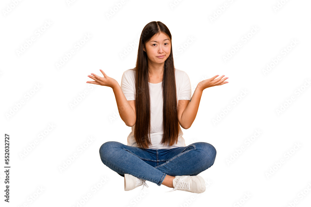 Young asian woman sitting on the floor cutout isolated doubting and shrugging shoulders in questioning gesture.