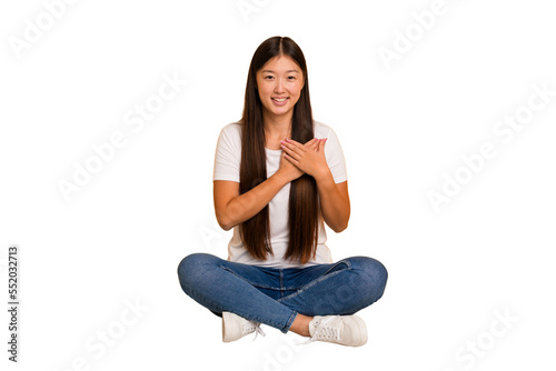 Young asian woman sitting on the floor cutout isolated has friendly expression, pressing palm to chest. Love concept. © Asier