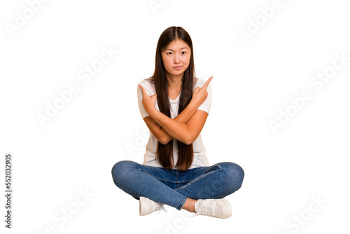 Young asian woman sitting on the floor cutout isolated points sideways, is trying to choose between two options.