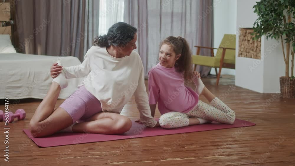 Young Hispanic Woman teach doing stretching exercises training her little cute daughter, sit floor in cozy living room perform in front of open laptop. Happy activity in family concept.