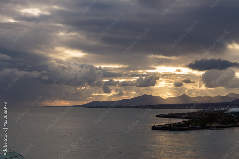 Incredible sunset on the beach. Storm clouds in the sky. Silhouette of mountains in the background. Golden sunbeams in the clouds. Beach in the dark. Lanzarote, Canary Islands, Spain.