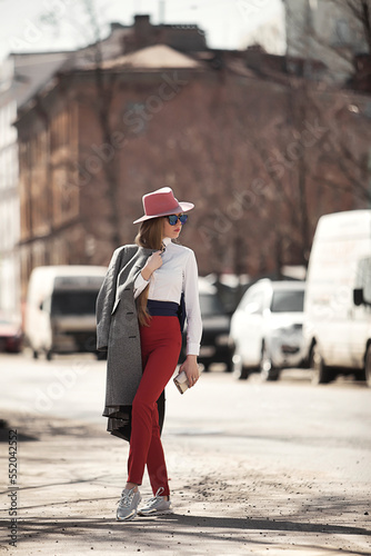 Stylish woman model in sunglasses walking in urban street wearing white shirt and red hat, looking away. Fashionable confident lady posing outdoors, lifestyle. Fashion style concept. Copy text space