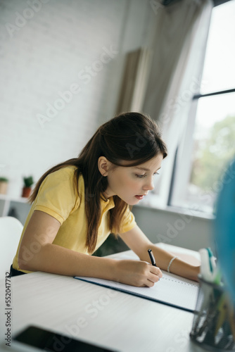 concentrated child writing in notebook during homeschooling on blurred foreground