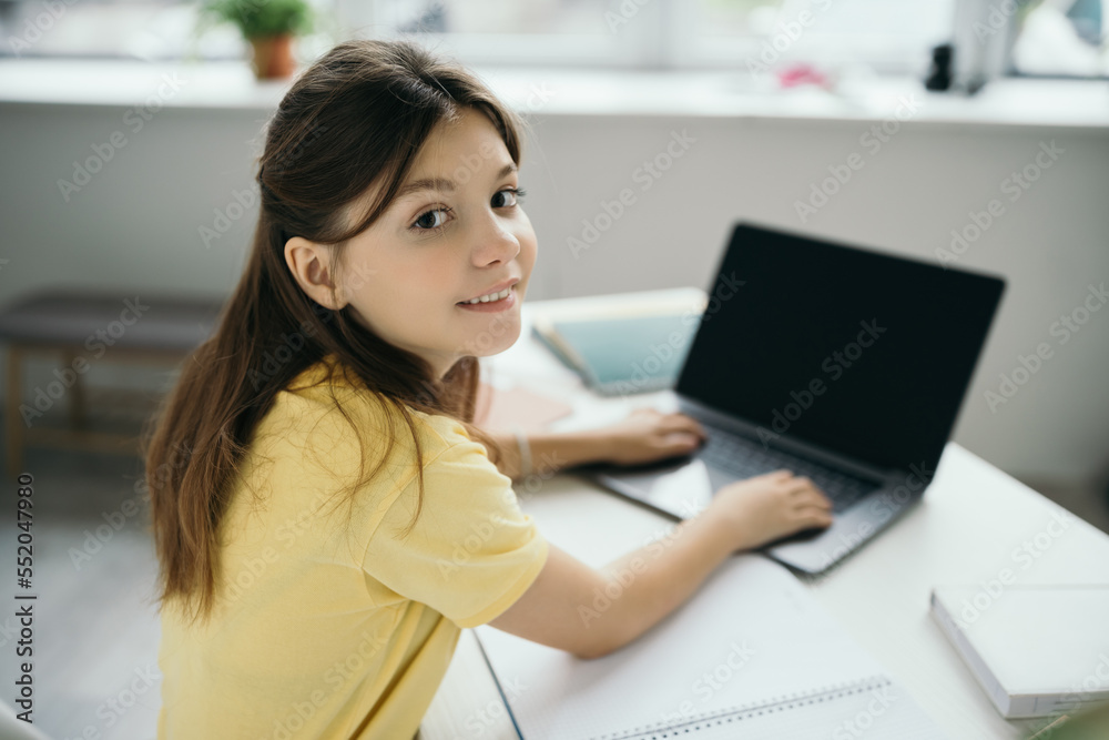 brunette girl smiling at camera and typing on blurred laptop while studying at home brunette girl smiling at camera and typing on blurred laptop while studying at home
