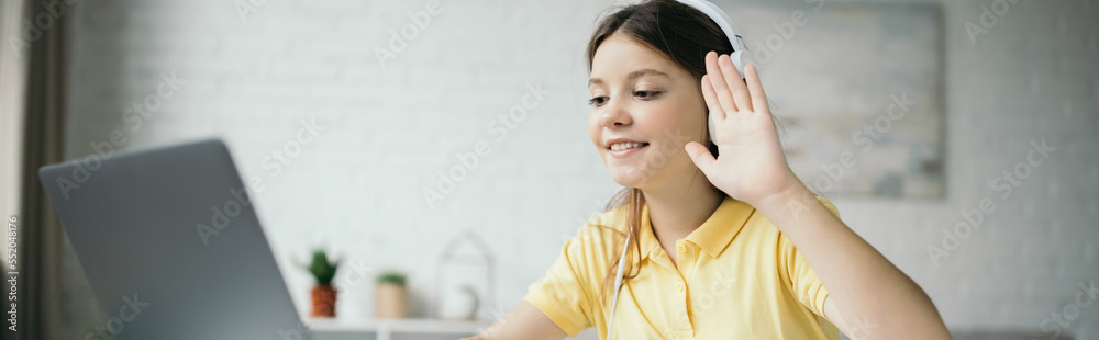 preteen girl in headphones waving hand and smiling during video call on ...