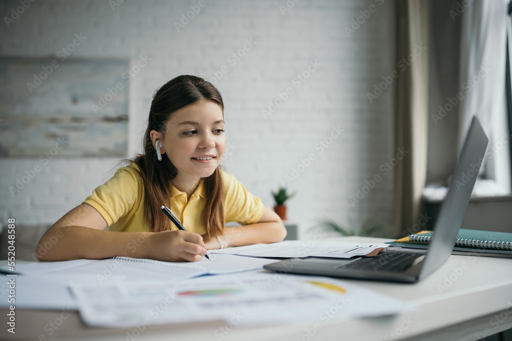 smiling girl in wireless earphone holding pen and looking at laptop at home smiling girl in wireless earphone holding pen and looking at laptop at home