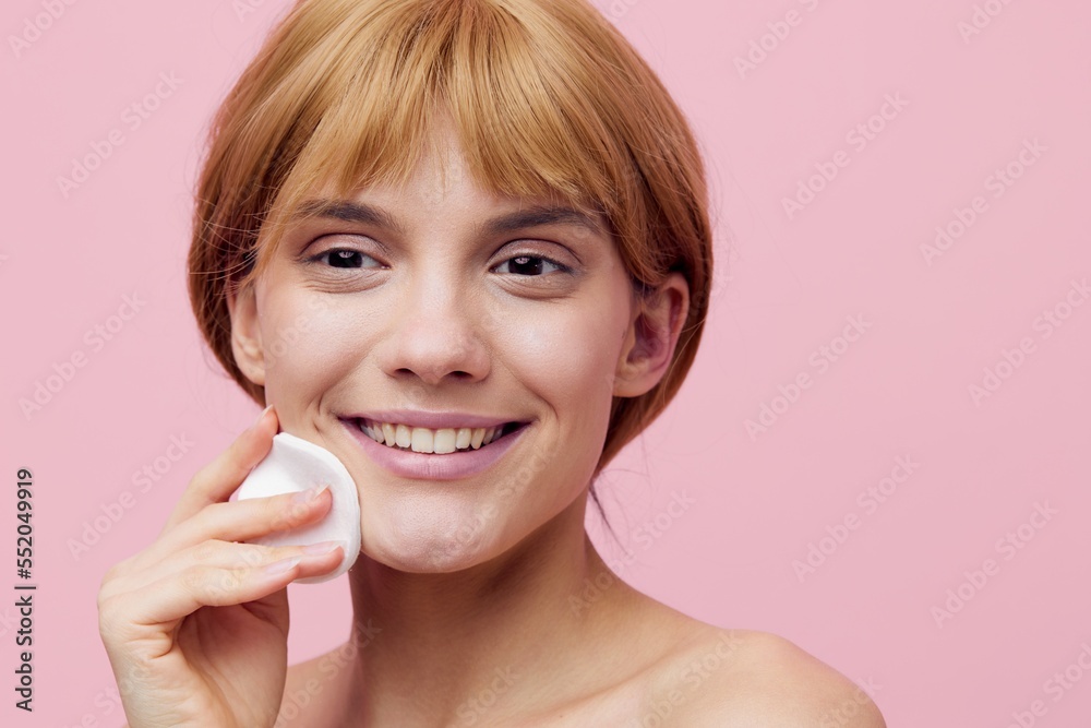a close horizontal photo on a pink background of a joyful woman with her hair pinned up from behind, smiling broadly at the camera, wipes her face with a cotton pad a close horizontal photo on a pink background of a joyful woman with her hair pinned up from behind, smiling broadly at the camera, wipes her face with a cotton pad