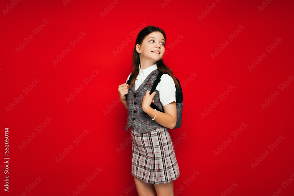 smiling girl in plaid clothes standing with backpack and looking back isolated on red, banner smiling girl in plaid clothes standing with backpack and looking back isolated on red, banner