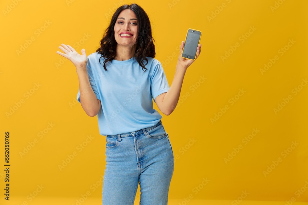 Woman blogger with a phone in her hands in a blue t-shirt and jeans on a yellow background smile signs gestures symbols, online communication and video call, copy space, free background
