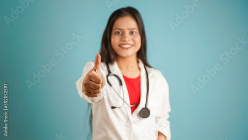 Portrait of a young female doctor gesturing thumbs up  Cheerful Asian Indian woman doctor in apron and stethoscope isolated over blue studio background