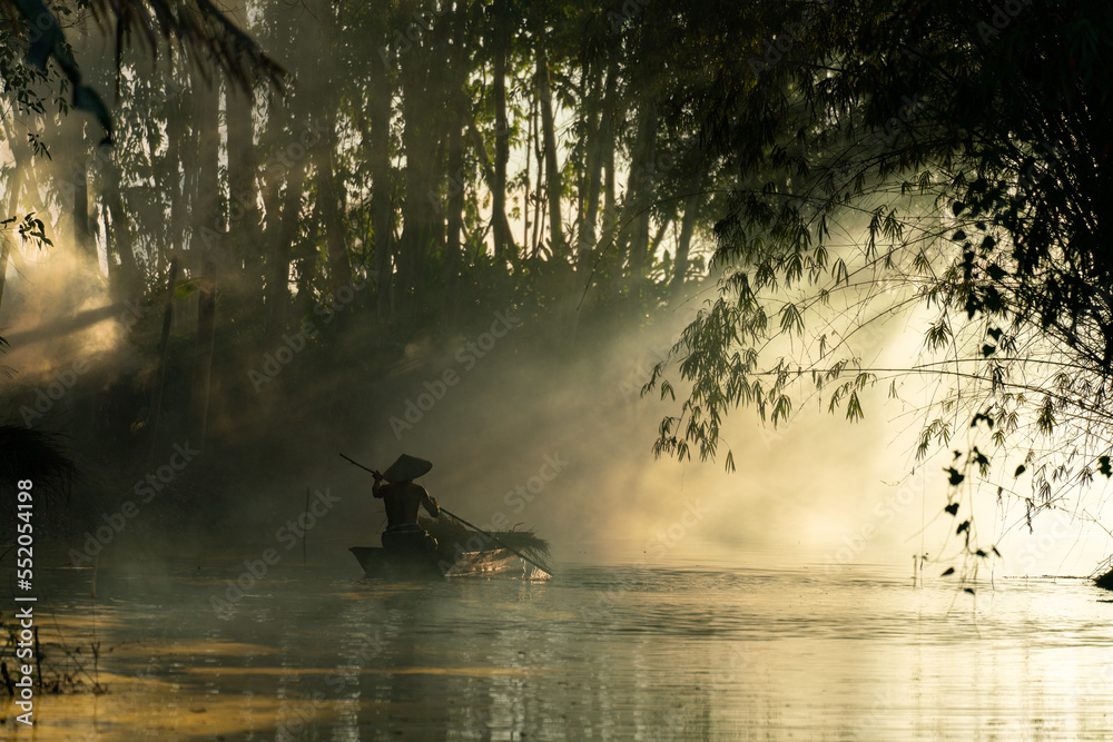 Photo of a local asian old man male boatman wearing conical hat rowing a small wooden boat across a small river during sunset time in a bamboo forest to deliver some dry grasses as animal feeds. Photo of a local asian old man male boatman wearing conical hat rowing a small wooden boat across a small river during sunset time in a bamboo forest to deliver some dry grasses as animal feeds.