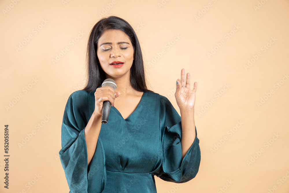 Beautiful young indian girl singer holding mic singing song isolated over beige studio background. Beautiful young indian girl singer holding mic singing song isolated over beige studio background.