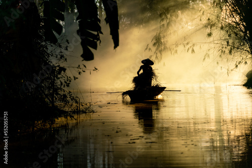 Photo of a local asian old man male boatman wearing conical hat rowing a small wooden boat across a small river during sunset time in a bamboo forest to deliver some dry grasses as animal feeds. 