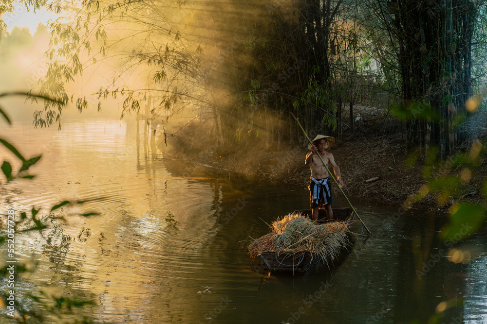 Φωτογραφία Photo of a local asian old man male boatman wearing conical hat rowing a small wooden boat across a small river during sunset time in a bamboo forest to deliver some dry grasses as animal feeds
